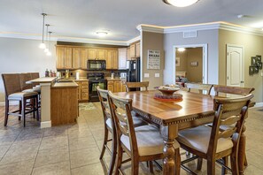 A modern kitchen with wooden cabinets, a central island, and a dining area with a wooden table and chairs. - Lakefront Patio, Pool Access! Lands End Condo (Osage Beach)