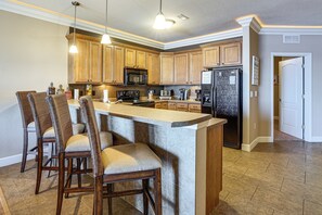 A kitchen with wooden cabinets, a central island with bar stools, a refrigerator, and a microwave. - Lakefront Patio, Pool Access! Lands End Condo (Osage Beach)