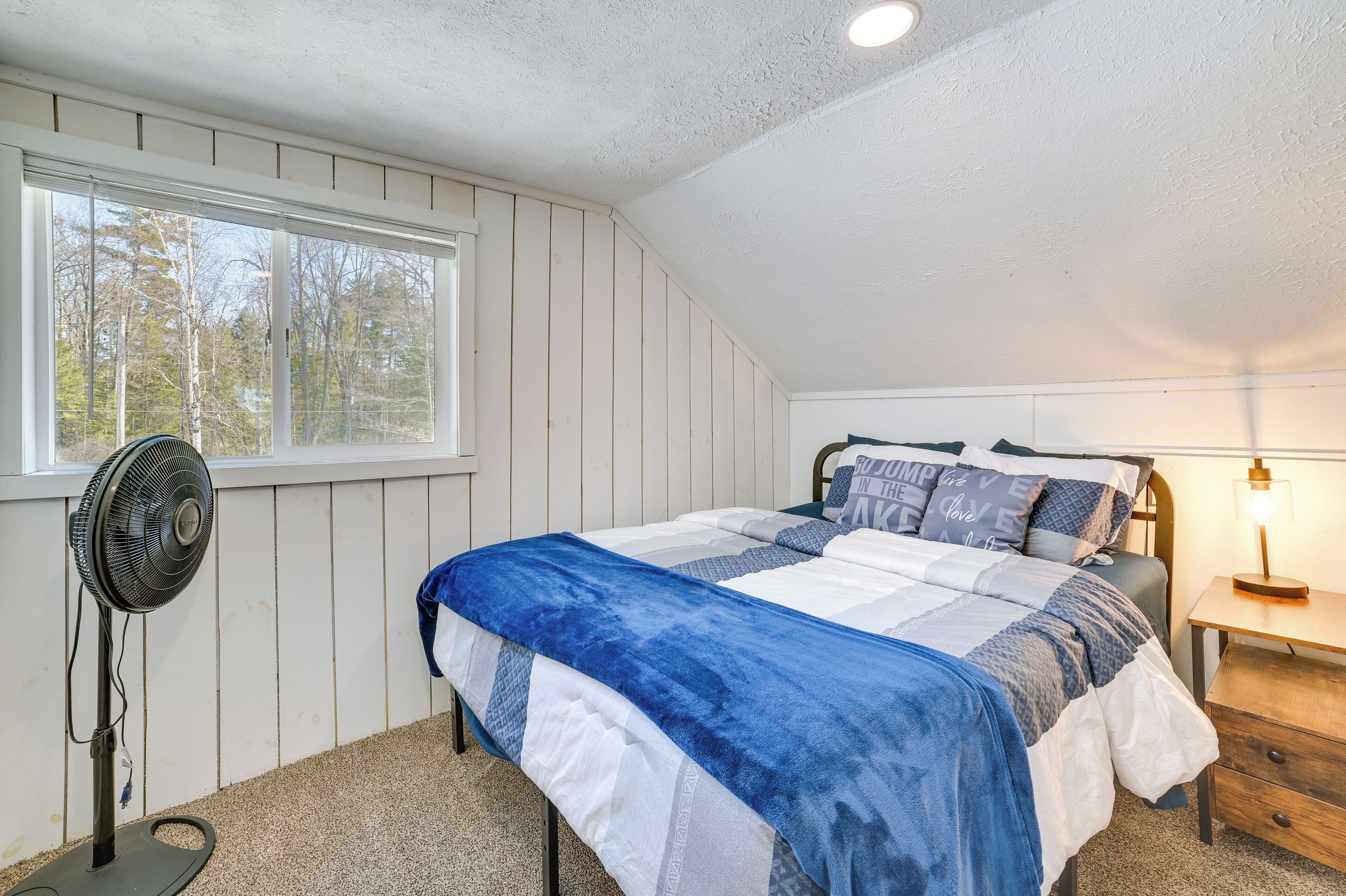 Una camera da letto con un letto, una ventola da terra, un comodino con una lampada e una finestra vista alberi.