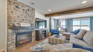 A cozy living room with a stone fireplace, a flat-screen TV, a comfortable sofa, and a large window offering a view. - Cozy Waterfront Hale Cottage on Long Lake! (Hale)