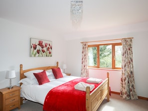 Une chambre avec un lit en bois, une literie blanche, un plaid rouge, une table de chevet avec une lampe, un tableau représentant des fleurs et une fenêtre avec des rideaux.