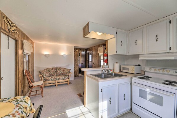 A compact kitchen with white cabinets, a built-in microwave, and a stove. There's a wooden chair and a floral sofa in the adjacent living area. - Pet-friendly Cabin w/ Dock: Fish & Explore! (Oriental)