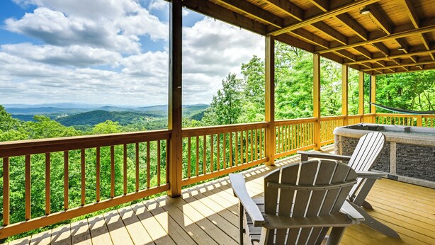 A wooden deck with Adirondack chairs and a stone fireplace, overlooking a lush forest and distant mountains. - Hot Tub & Game Room: Mountaintop Cabin in Murphy! (Murphy)