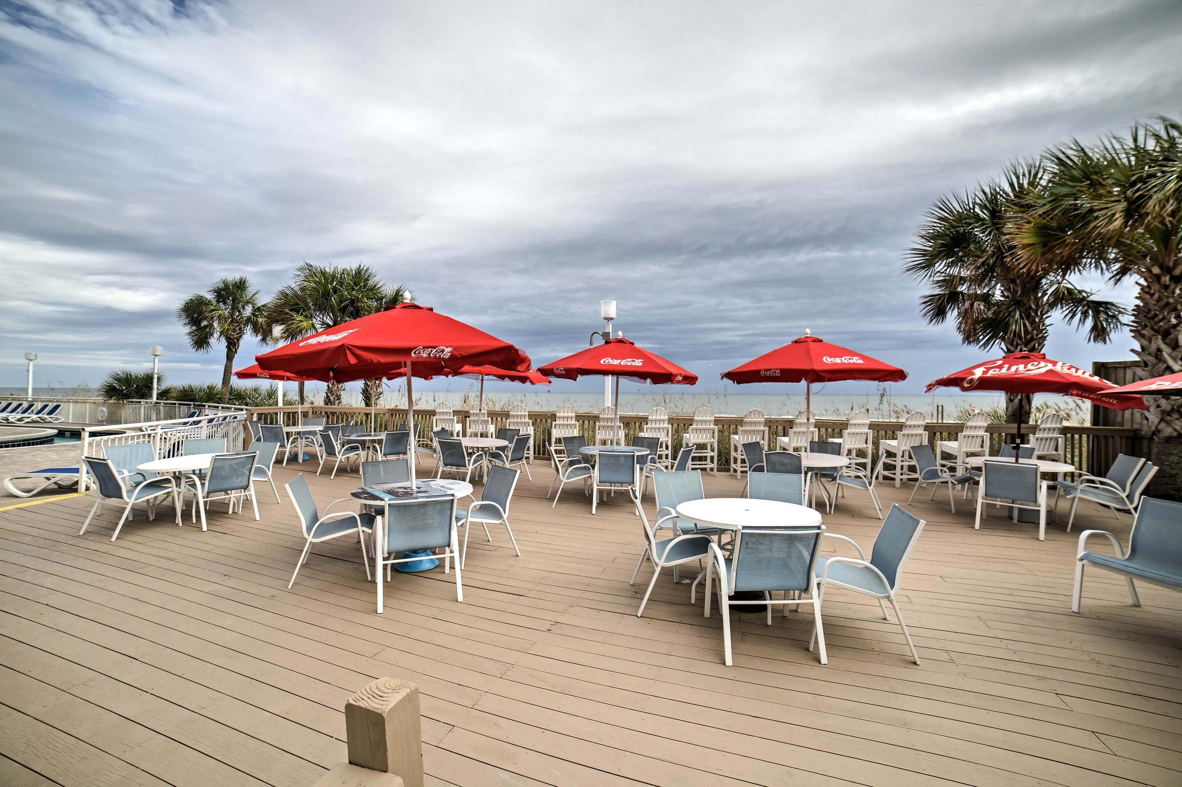 An outdoor deck with tables and chairs, red umbrellas, and a view of the ocean.