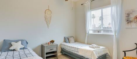 Un dormitorio con dos camas, un ventilador de techo, una ventana con cortinas, una mesita de noche y una silla.