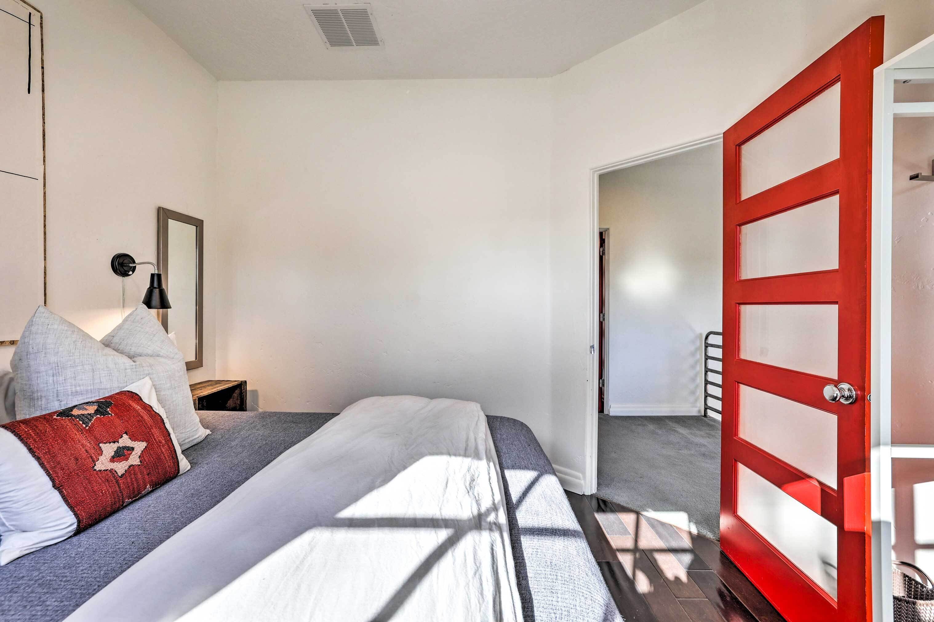 A bedroom with a bed, a nightstand, a lamp, a red door, and a hallway.