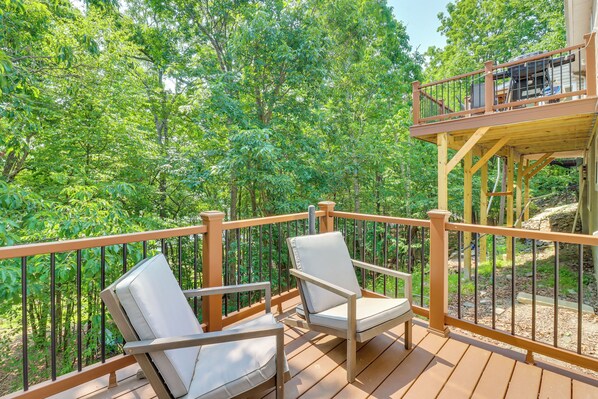 A wooden deck with two white cushioned chairs, surrounded by a metal railing, overlooking a lush green forest. - Gas Grill, Lake Access: Scenic Poconos Retreat! (East Stroudsburg)