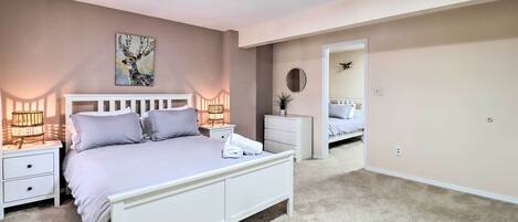 Un dormitorio con cama blanca, dos mesitas de noche con lámparas, un espejo y una puerta que da a otra habitación.