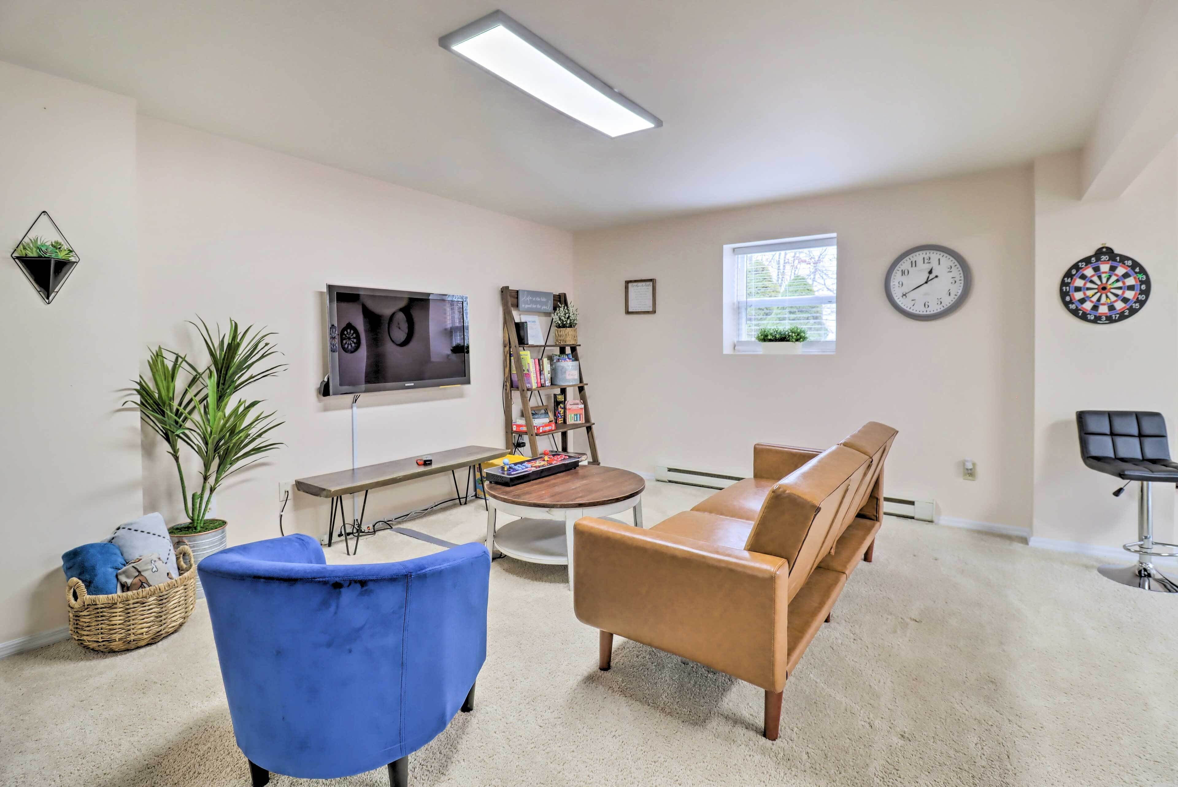 A living room with a television, a bookshelf, a coffee table, a blue chair, a brown sofa, a window, and a dartboard.
