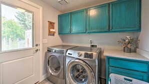 A laundry room with a front-loading washer and dryer, teal cabinets, and a window with blinds. - Idyllic Nampa Family Home With Hot Tub & Fire Pit! (Nampa)