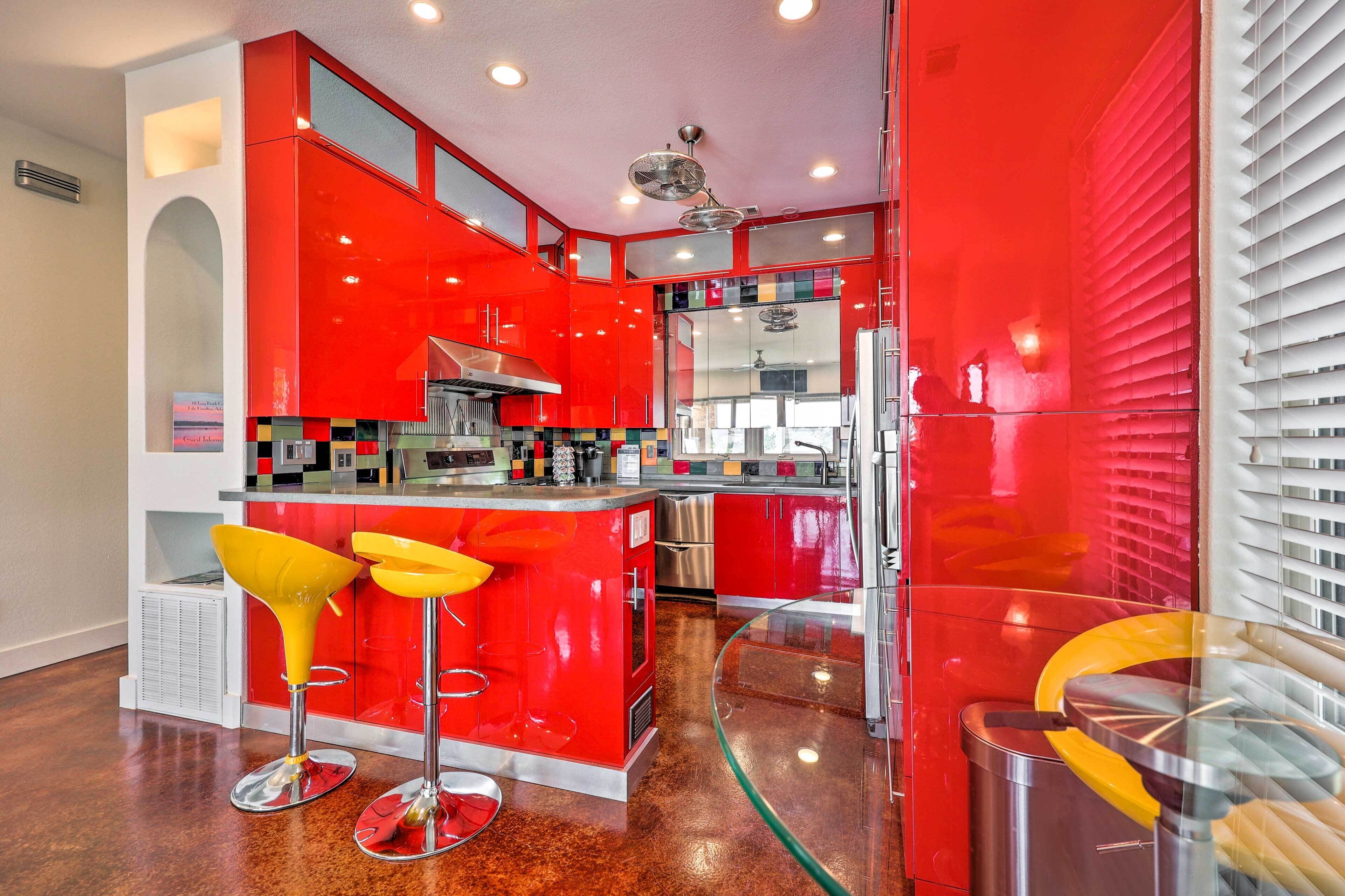 A modern kitchen with red cabinetry, stainless steel appliances, and a glass island with yellow bar stools.