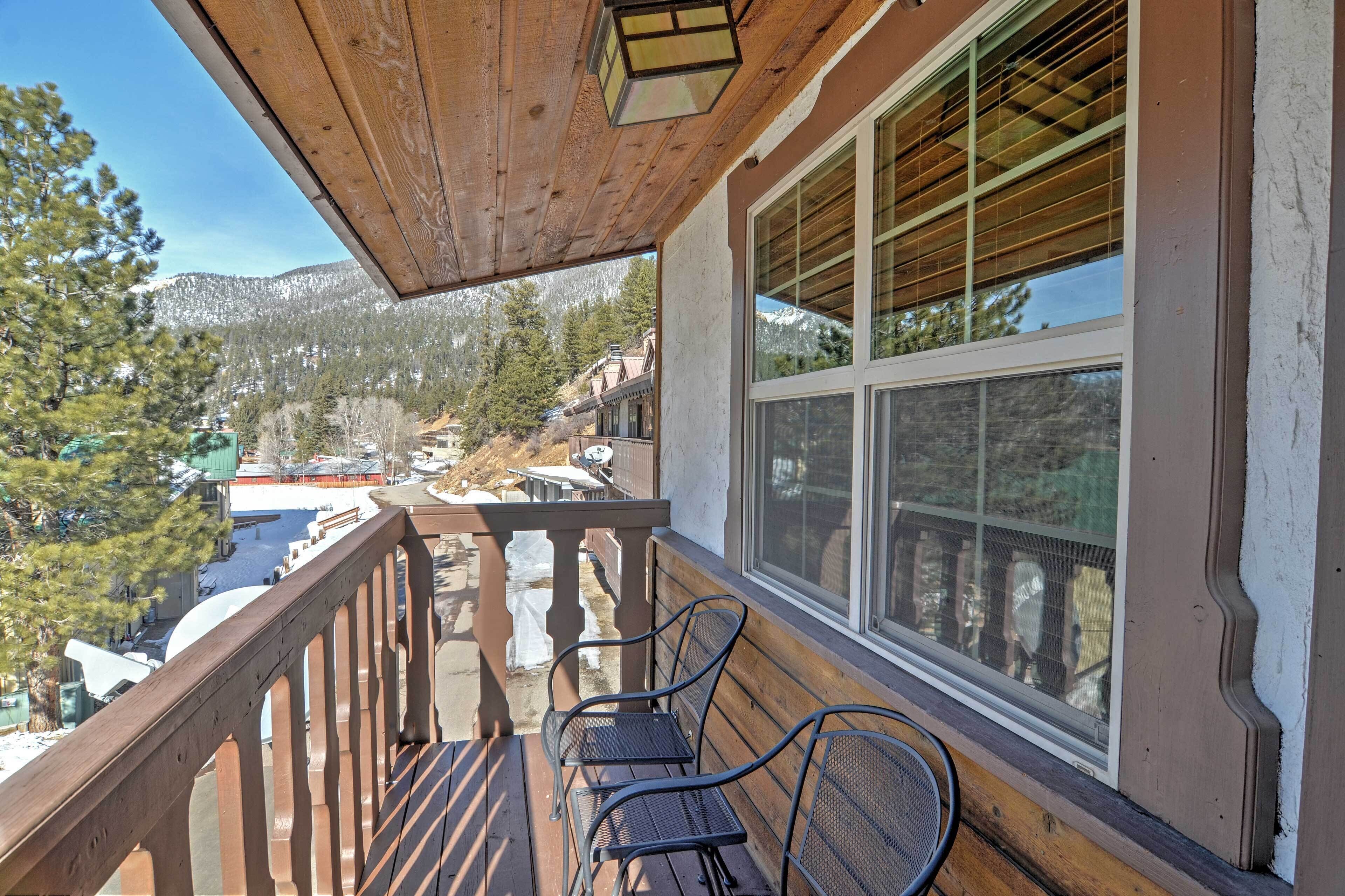 A wooden balcony with metal chairs, a view of a snowy mountain, and a building with large windows.