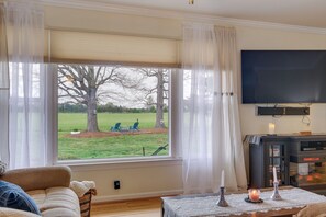 A living room with a large window, a television, a fireplace, and a table with candles. - Deck & Pasture Views! Bealeton Farmhouse Escape (Bealeton)