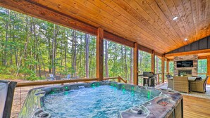 Un jacuzzi con chimenea de piedra y mobiliario exterior, rodeado por una terraza de madera y vistas al bosque.