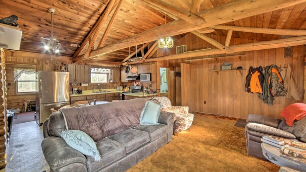 A log cabin interior with a wooden ceiling, a sofa, a dining area, and a kitchen. - Cornell Home w/ Fire Pit & Hunting Access! (Cornell)