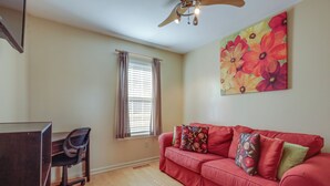 A living room with a red sofa, a desk, and a chair. There is a window with curtains and a ceiling fan. - Tranquil Delmar Home ~ 7 Mi to Salisbury! (Delmar)