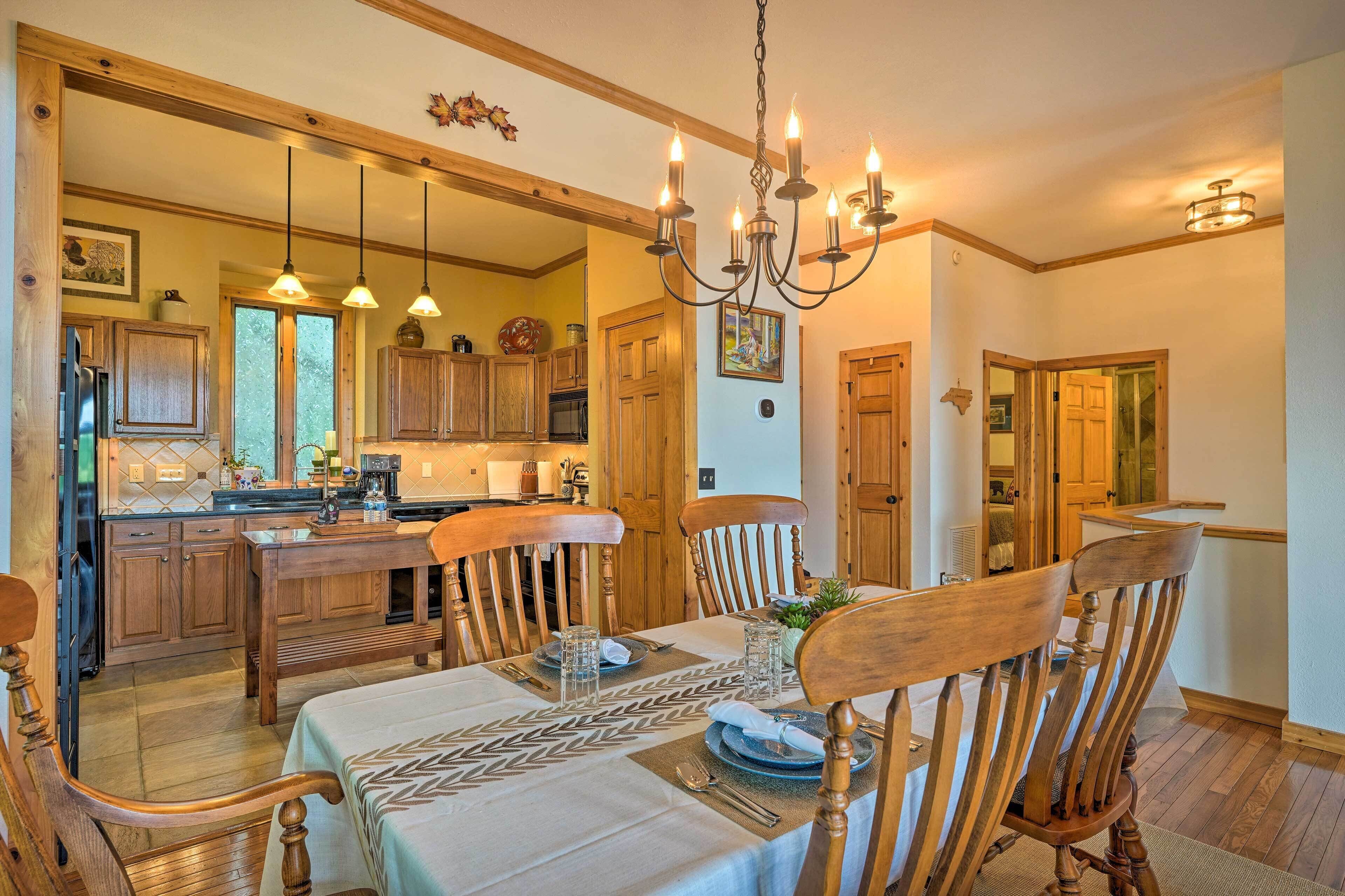 Un comedor con una mesa de madera dispuesta para cuatro personas, rodeada de sillas de madera. Un candelabro cuelga sobre la mesa, y la habitación tiene suelos y paredes de madera.