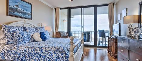 Une chambre avec un grand lit, un ventilateur de plafond, un tableau, un balcon avec vue et une commode.