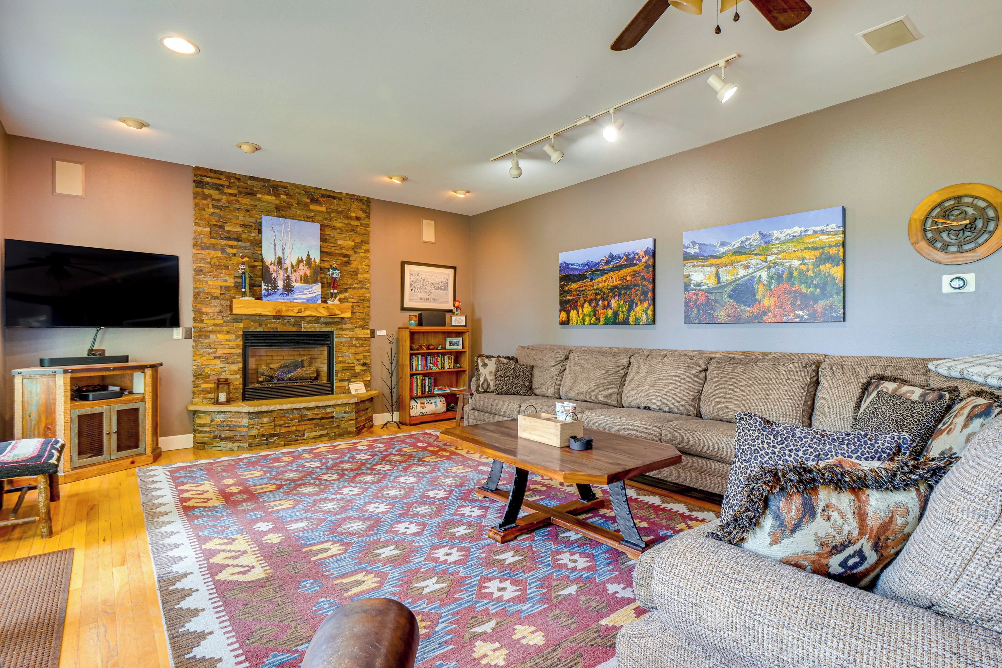 A cozy living room with a stone fireplace, a large flat-screen TV, a sofa, a coffee table, and a colorful rug.