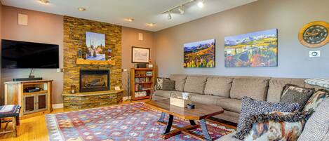 A cozy living room with a stone fireplace, a large flat-screen TV, a sofa, a coffee table, and a colorful rug.