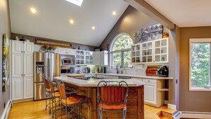 Une cuisine moderne avec des meubles blancs, un grand îlot central doté de tabourets, un four encastré et une fenêtre donnant sur la verdure.