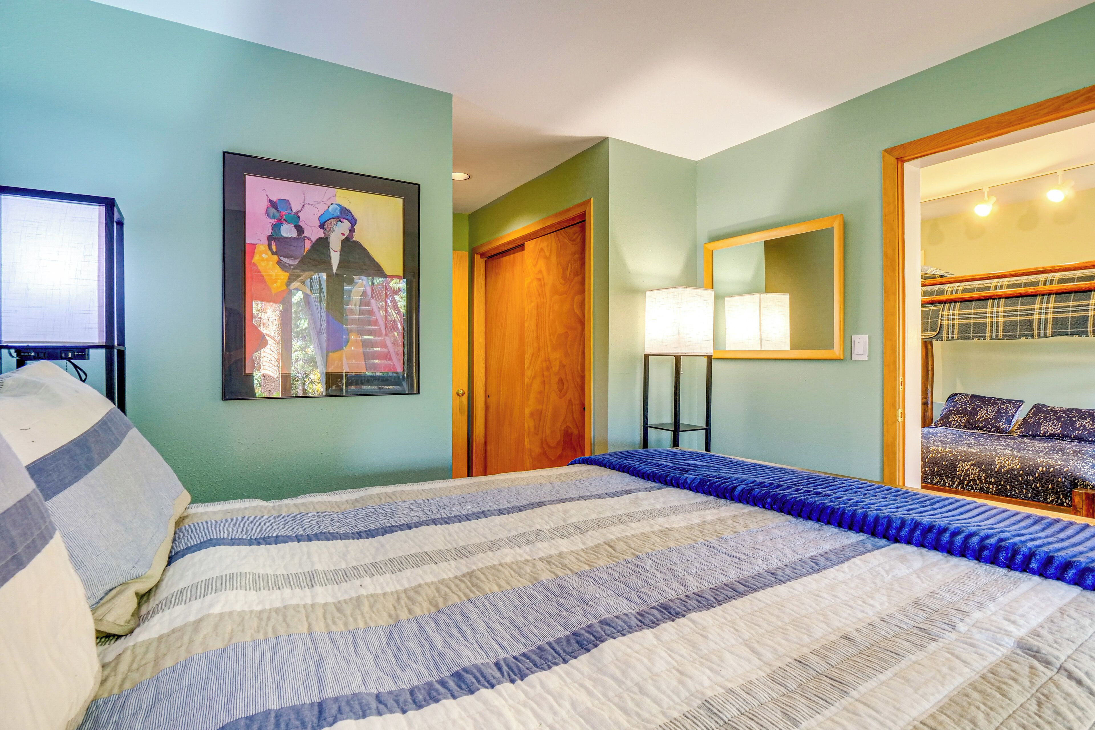 A bedroom with a bed, a wooden door, a framed painting, a bedside lamp, and a bunk bed in the adjacent room.
