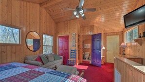 A wooden cabin interior with a bed, a sofa, a ceiling fan, a television, and a bathroom visible through an open door.