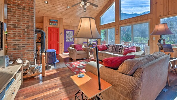 A cozy cabin interior with a fireplace, wooden floors, a ceiling fan, large windows, and a comfortable seating area.
