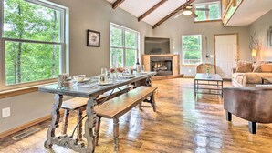 A spacious living room with a large wooden table, a fireplace, and large windows offering a view of the outdoors. - Hike & Zip-line: Red River Gorge Cabin w/ Hot Tub! (Zoe)