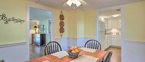 Une salle à manger avec une table en bois, quatre chaises, un ventilateur de plafond et un panier de fruits posé sur la table.
