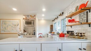 Una cocina moderna con armarios blancos, microondas, hervidor de agua y cafetera. Hay estantes con objetos decorativos y un letrero que dice 'Un perro es la única criatura que te ama más a sí misma'.