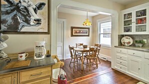 A kitchen with wooden cabinets, a coffee maker, a dining table with chairs, and a painting of a cow.
