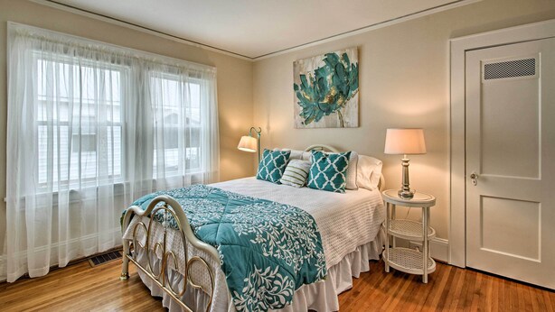 A bedroom with a bed, two bedside lamps, a window with sheer curtains, a door, and a wall-mounted artwork.