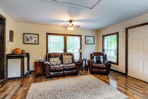 Un salon avec un canapé en cuir marron, un fauteuil assorti, une table basse en bois et un tapis à motifs. Il y a deux fenêtres avec des stores, un ventilateur de plafond et des tableaux encadrés accrochés aux murs.