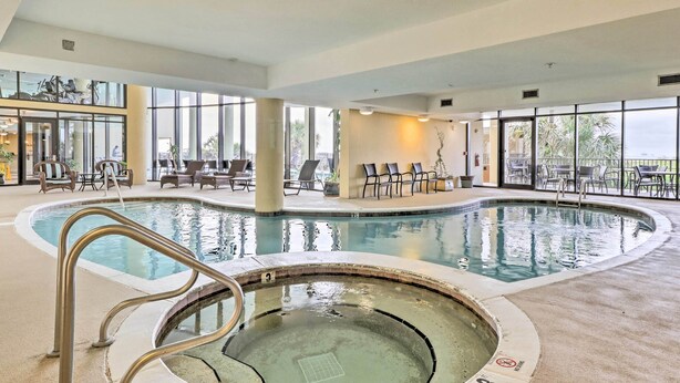 An indoor swimming pool with a hot tub, lounge chairs, and a view of the outdoors.
