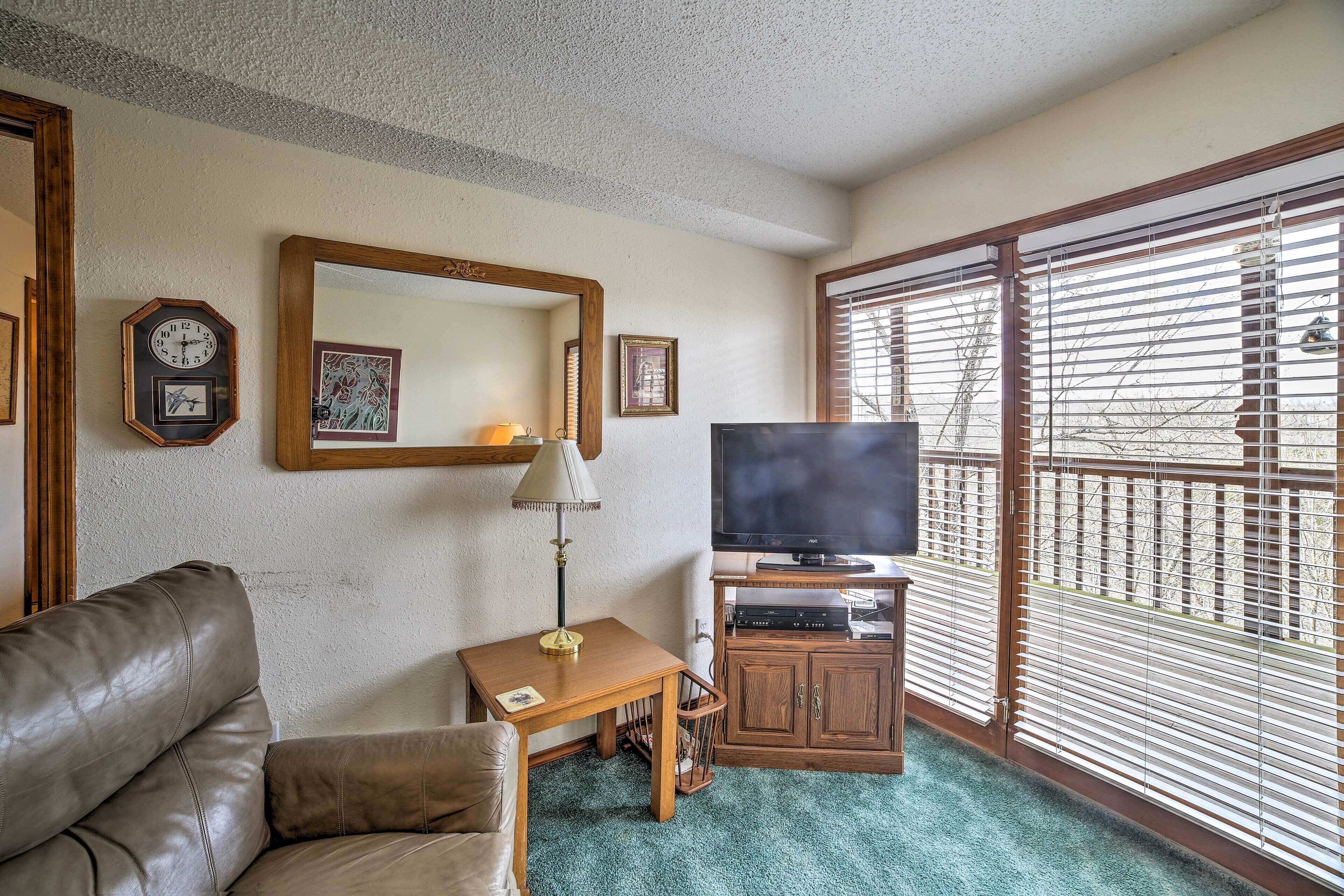 A living room with a leather sofa, a small table with a lamp, a television on a wooden stand, and a large window with blinds.