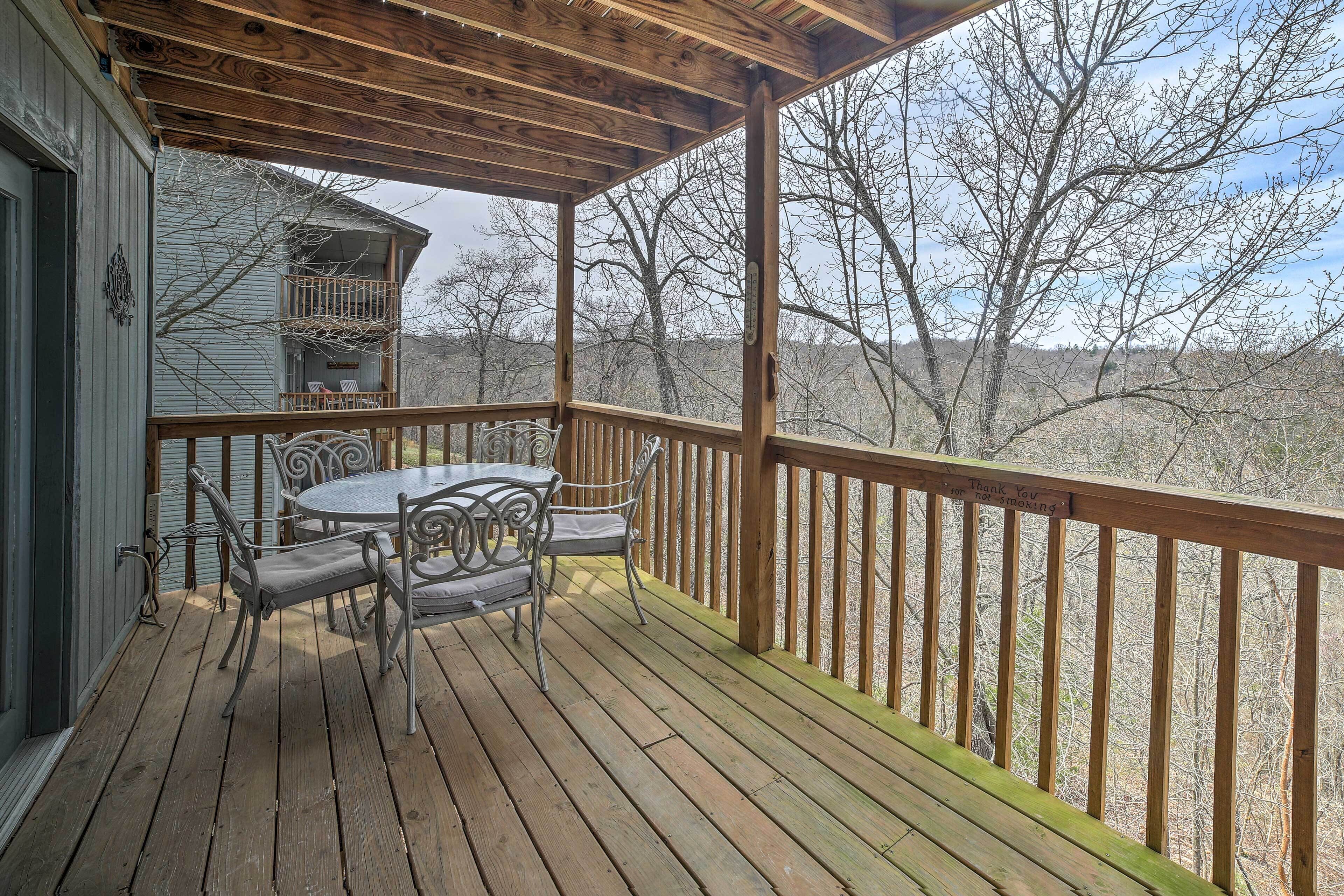Une terrasse en bois avec une table et des chaises, un espace abrité et une vue sur des arbres et une maison au loin.