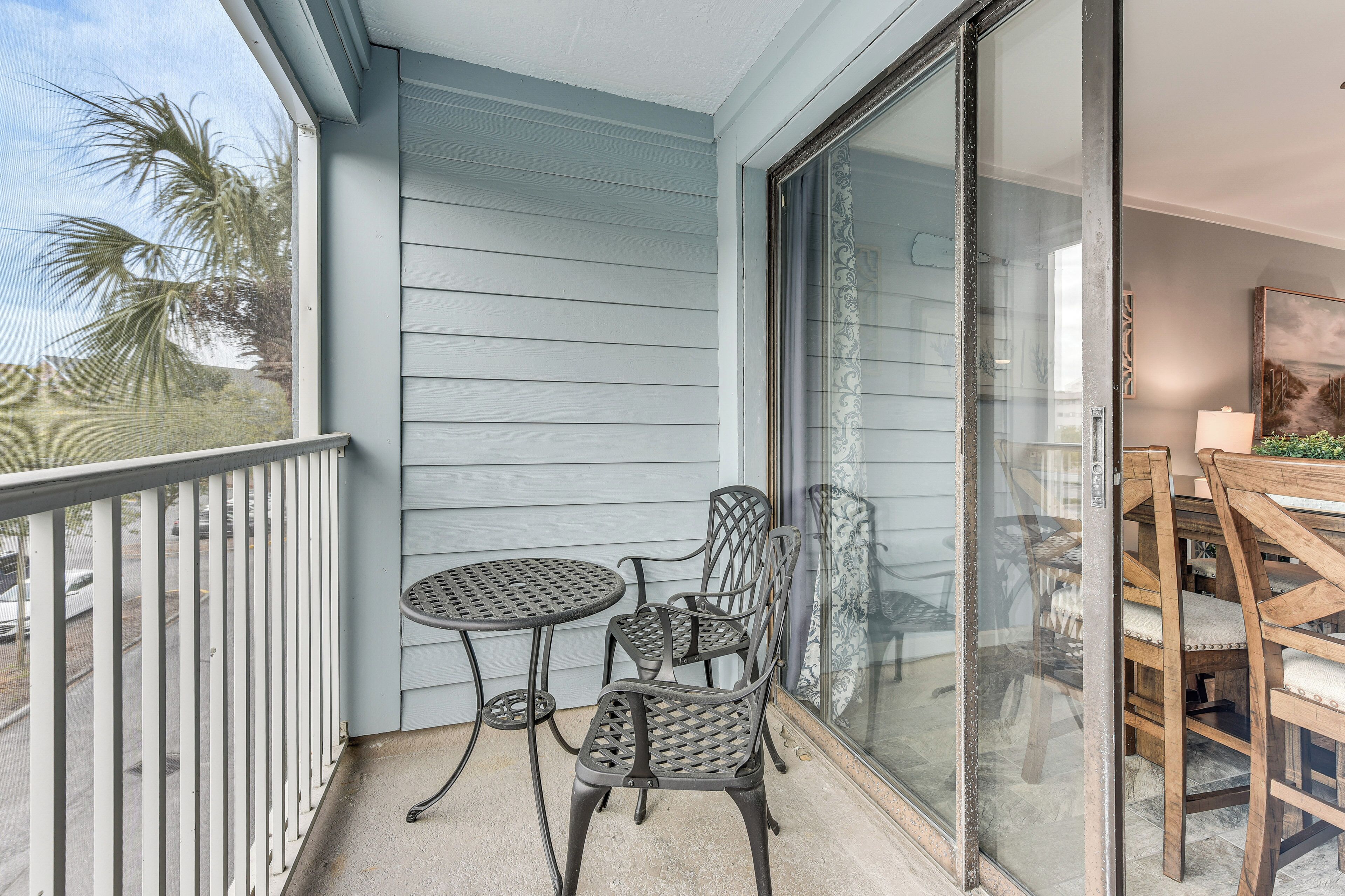 A balcony with a table and two chairs, a sliding glass door, and a view of palm trees.