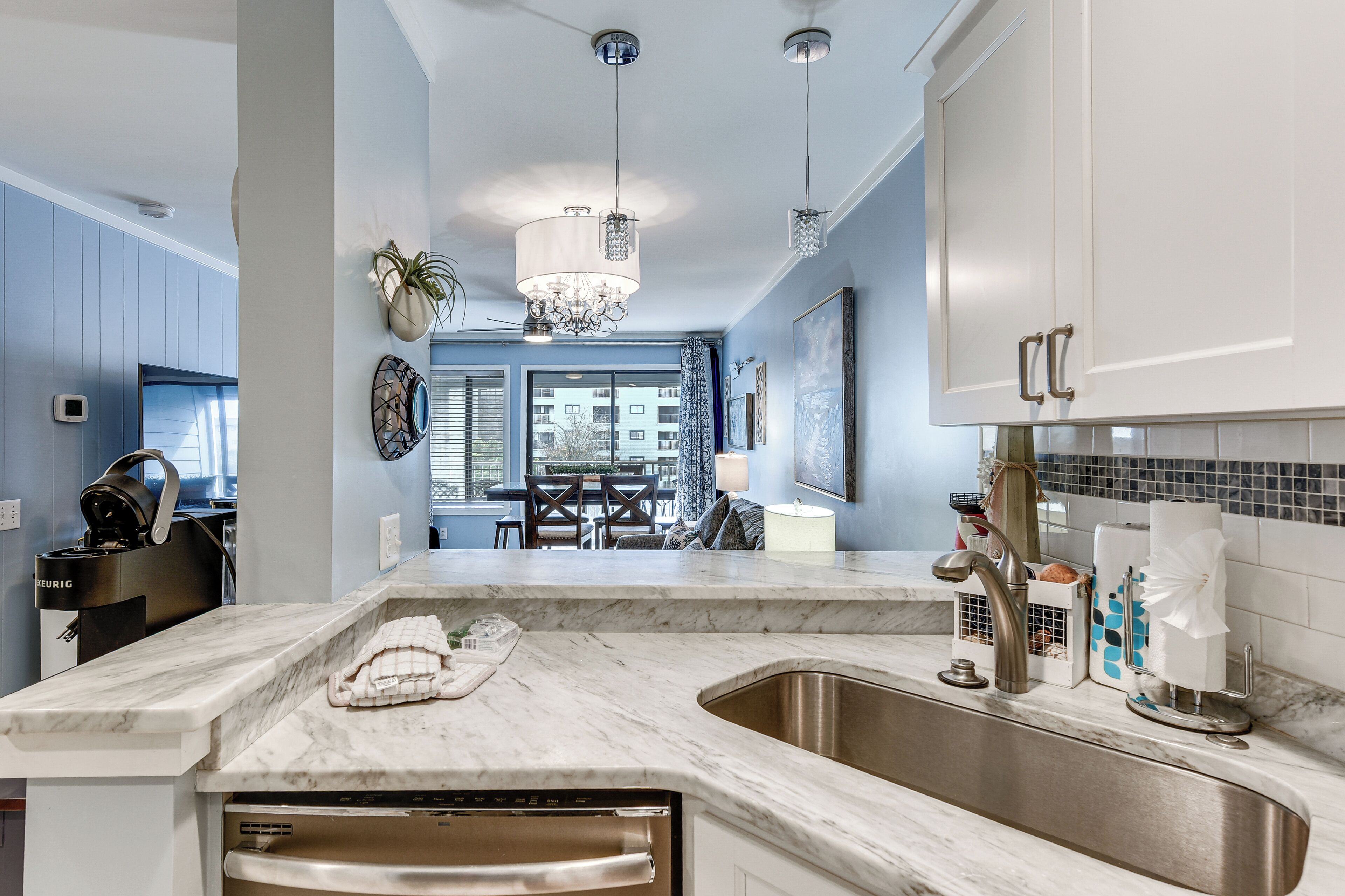 A modern kitchen with a marble countertop, stainless steel sink, and a built-in dishwasher. There is a dining area visible in the background with a table and chairs.