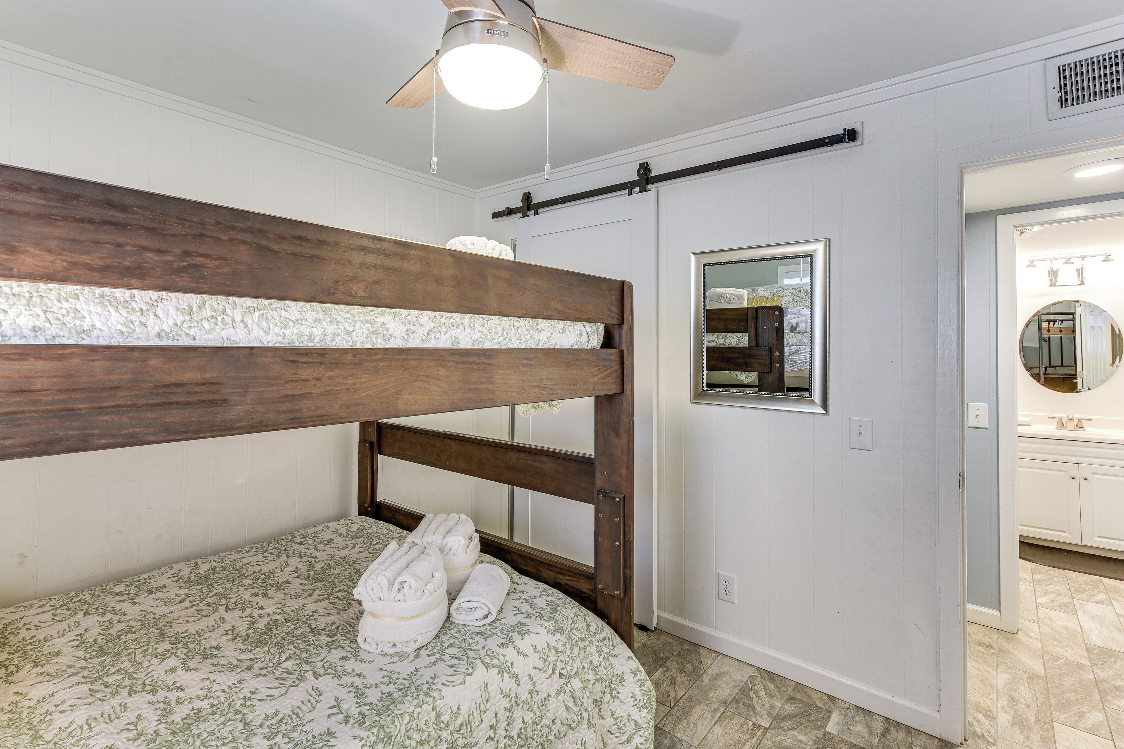 A bedroom with a bunk bed, a bedside table with towels, a ceiling fan, and a bathroom with a mirror and sink.