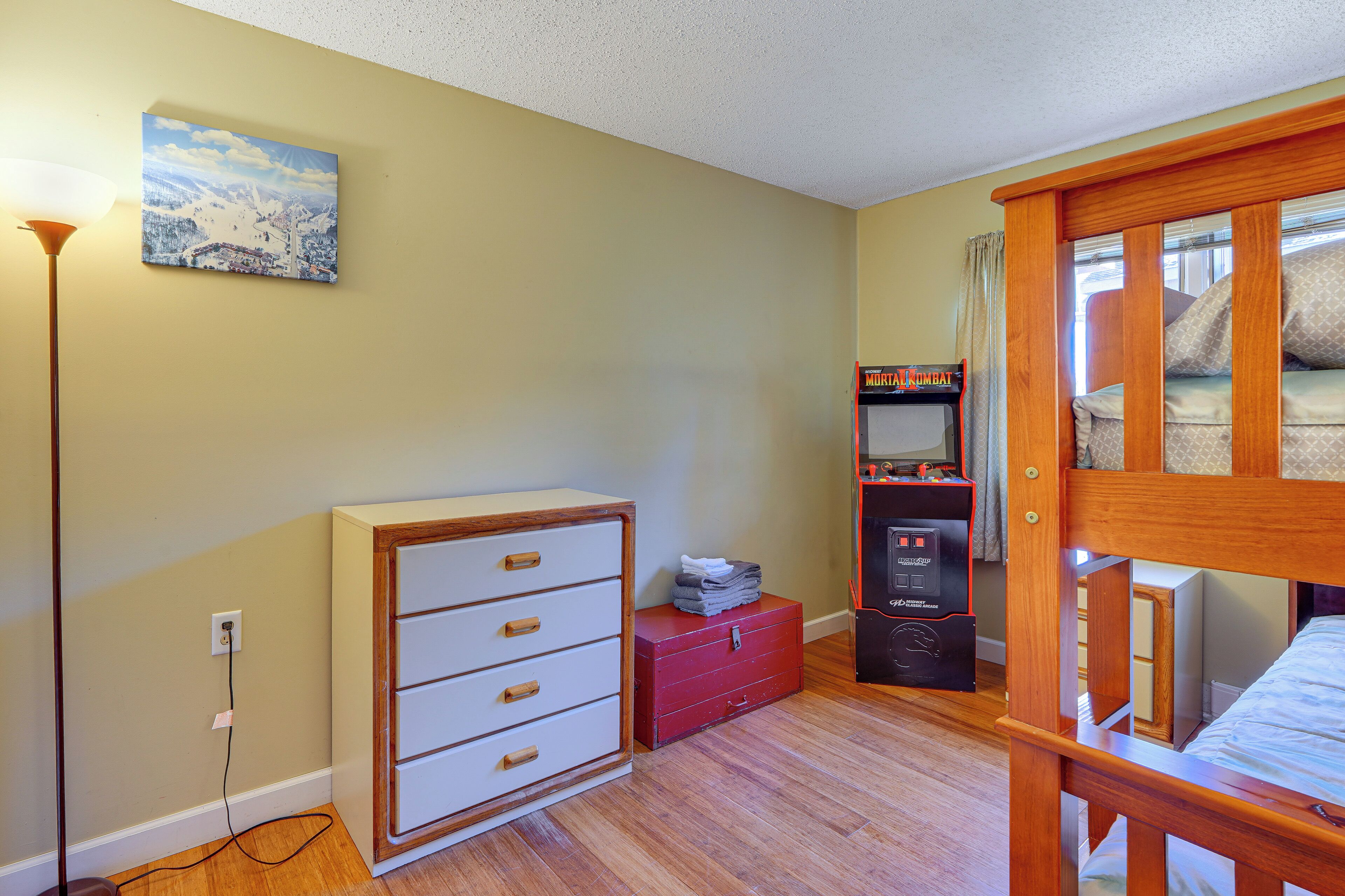 A bedroom with a bunk bed, a wooden dresser, a red chest of drawers, a painting on the wall, and a gaming console.