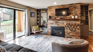A living room with a fireplace, a television mounted on a stone wall, and a sliding glass door leading to a balcony. - Golf & Adventure! Condo 2 Mi to Dtwn Ellicottville (Great Valley)