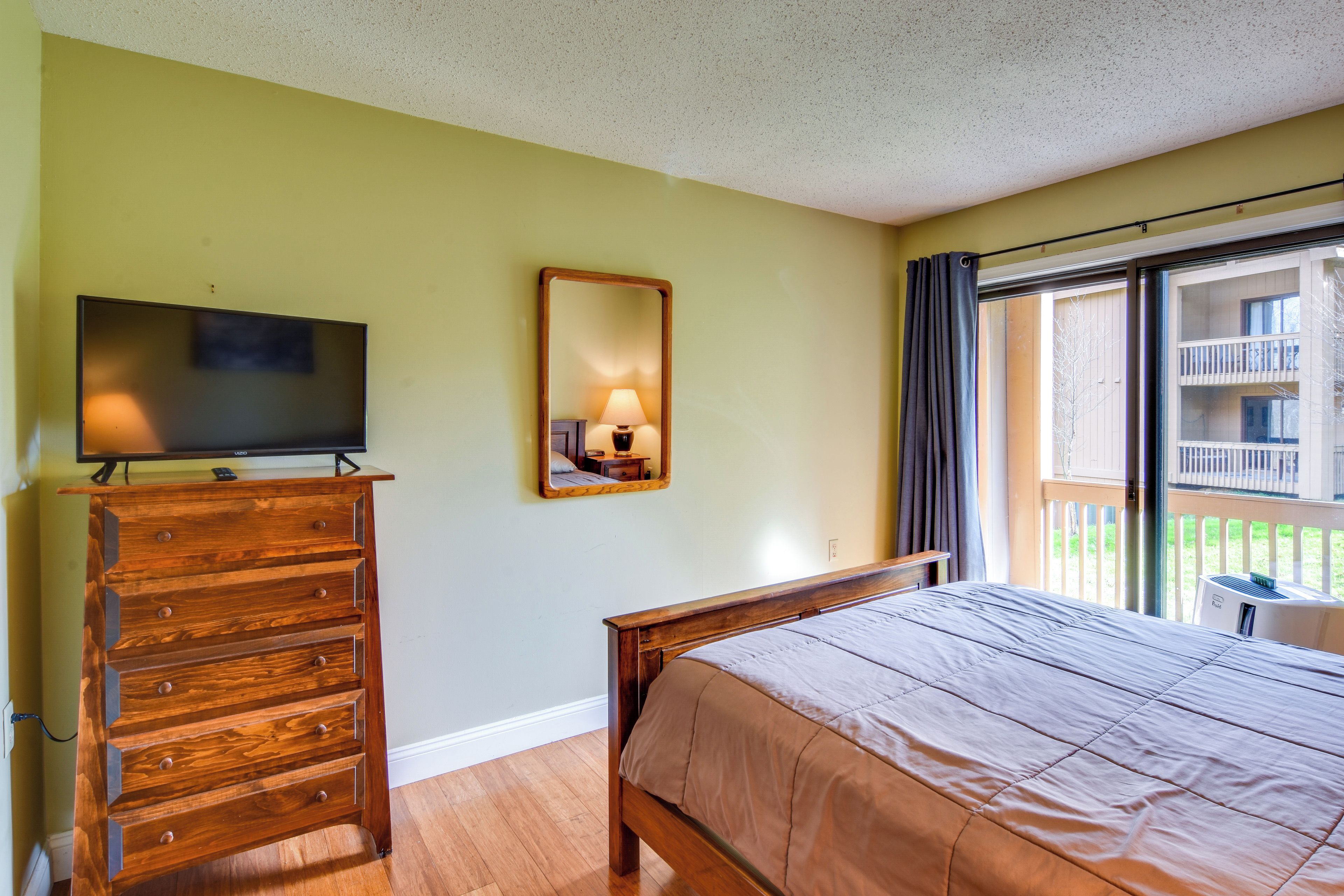 A hotel room with a bed, a wooden dresser, a flat-screen TV, a mirror, and a balcony with a view of the outside.