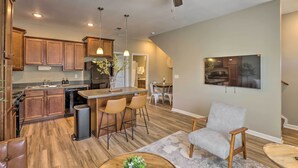 Une cuisine spacieuse avec des meubles en bois, un îlot central doté de tabourets de bar, un ventilateur de plafond, un éclairage encastré et un coin repas avec un téléviseur fixé au mur.