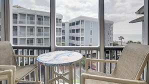 Een balkon met rieten stoelen en een ronde tafel, met uitzicht op een gebouw aan het strand met meerdere balkons.