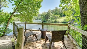 A wooden deck with wicker chairs and a round table overlooking a lake, surrounded by trees and a wooden fence. - The Bella Vista Lakeside Treehouse: Dock & Kayaks! (Bella Vista)