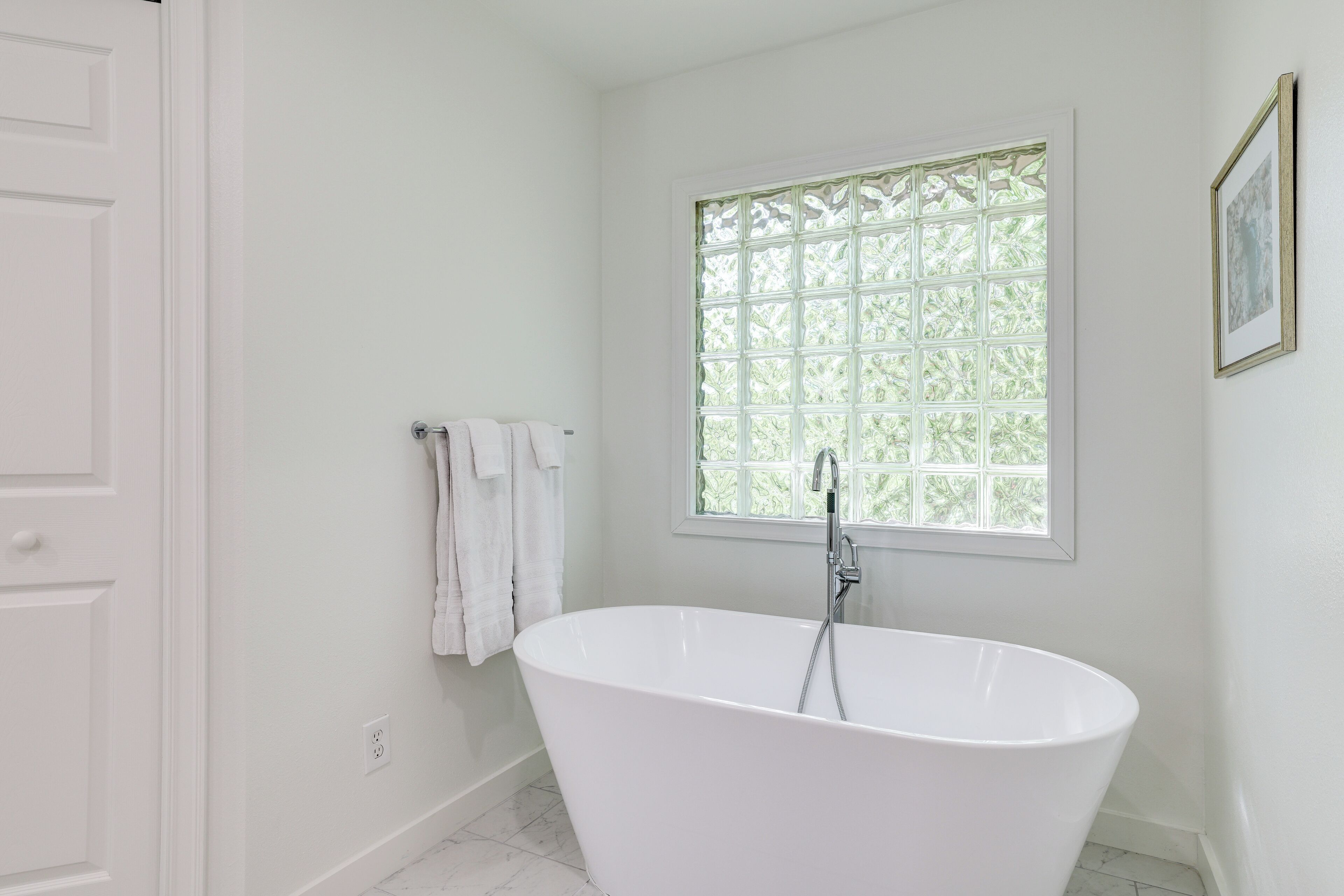 A white bathroom with a freestanding bathtub, a window with a grid pattern, and a framed picture on the wall.