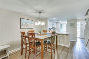 A dining area with a wooden table and chairs, a chandelier, and a kitchen in the background.