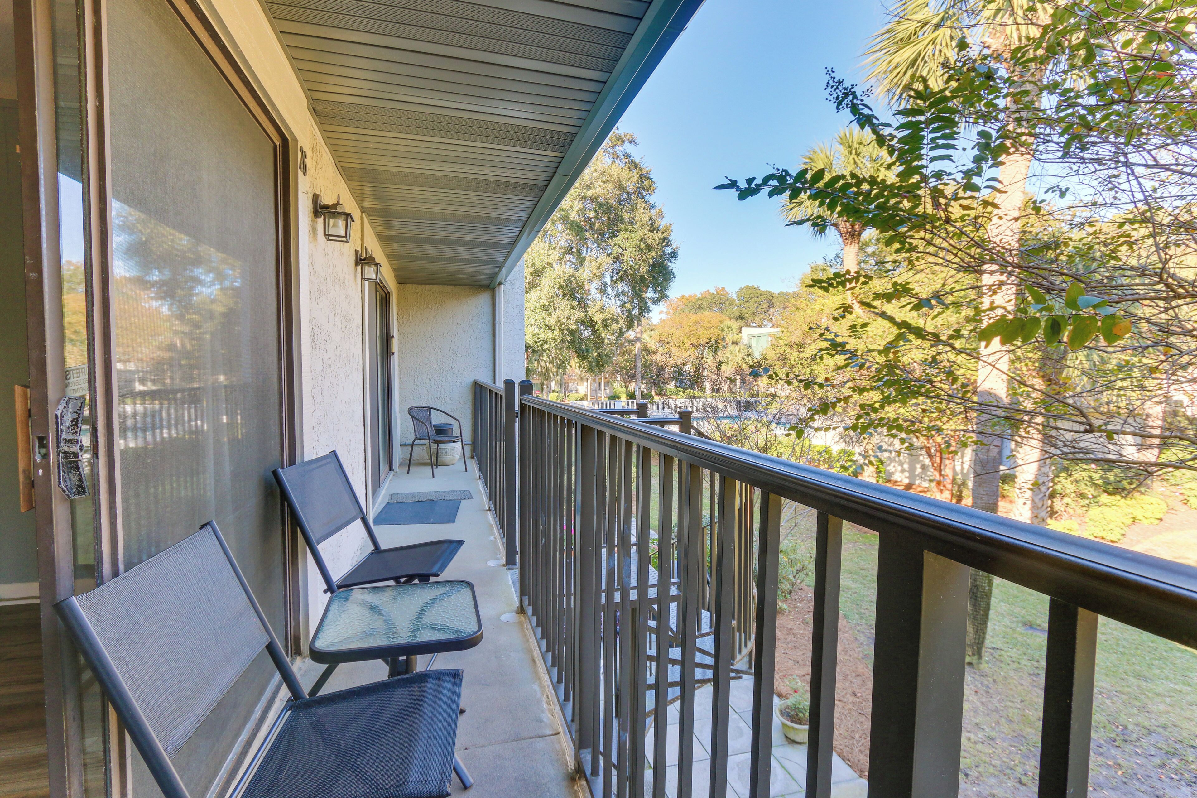 A balcony with two chairs and a table, overlooking a garden with trees and a clear sky.