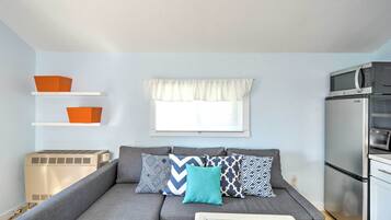 A compact living space with a grey sofa, patterned cushions, a window with white valance, a wall-mounted air conditioner, and a kitchenette with a microwave and refrigerator.